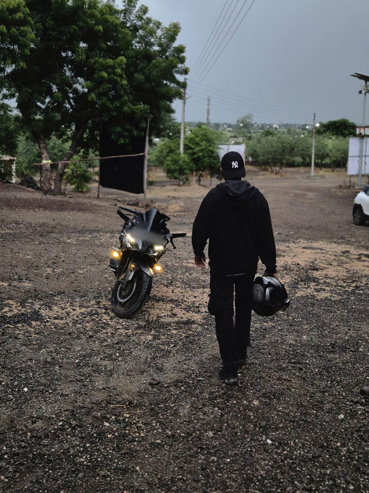 A rider in a black hoodie and cap walking toward a parked motorcycle with its lights on, under a stormy sky